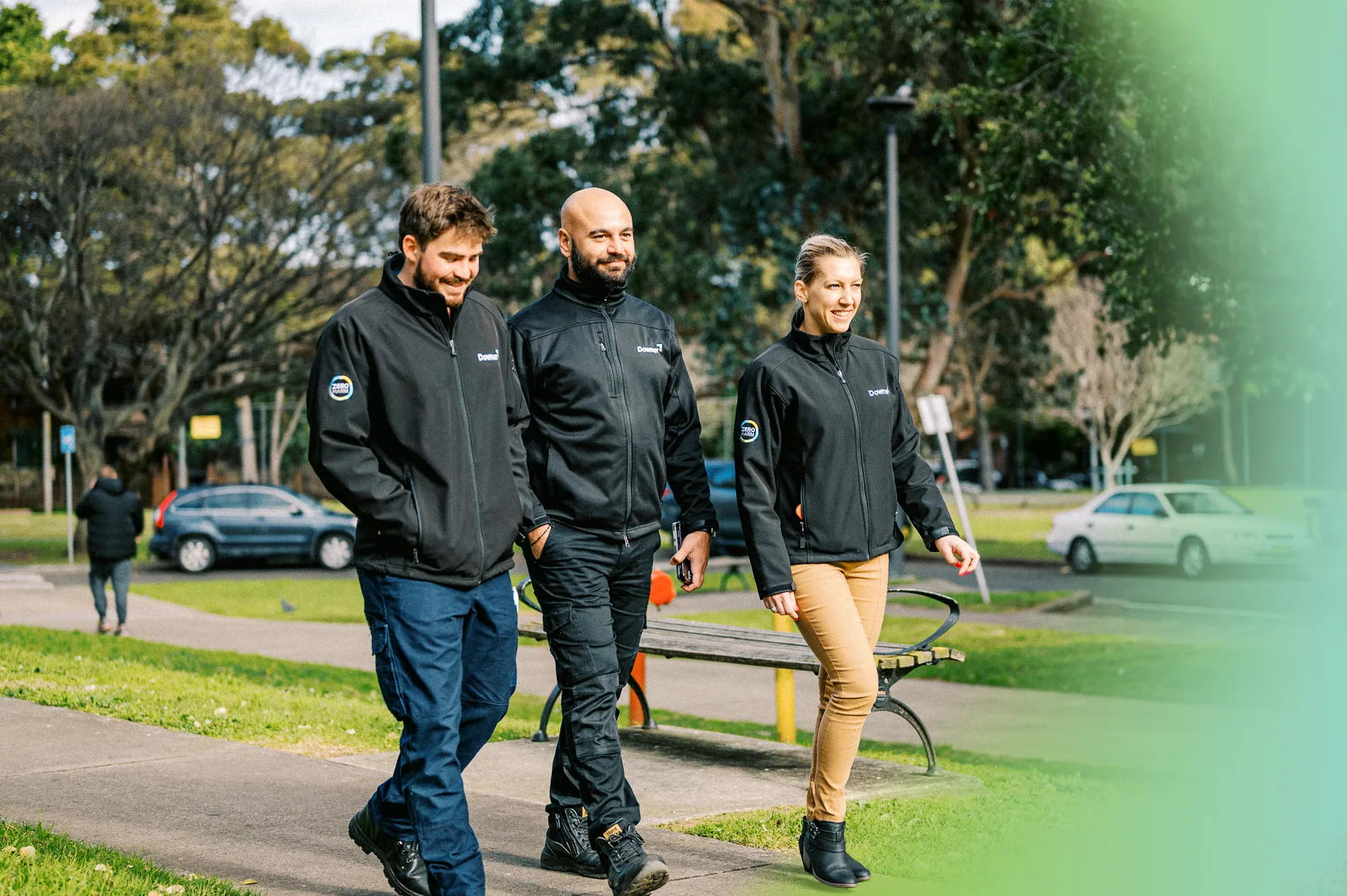 Three colleagues wearing black Downer-branded jackets walk and talk together on a paved path in an outdoor park setting. They appear to be in conversation against a backdrop of green trees and a soft-focus urban park environment.