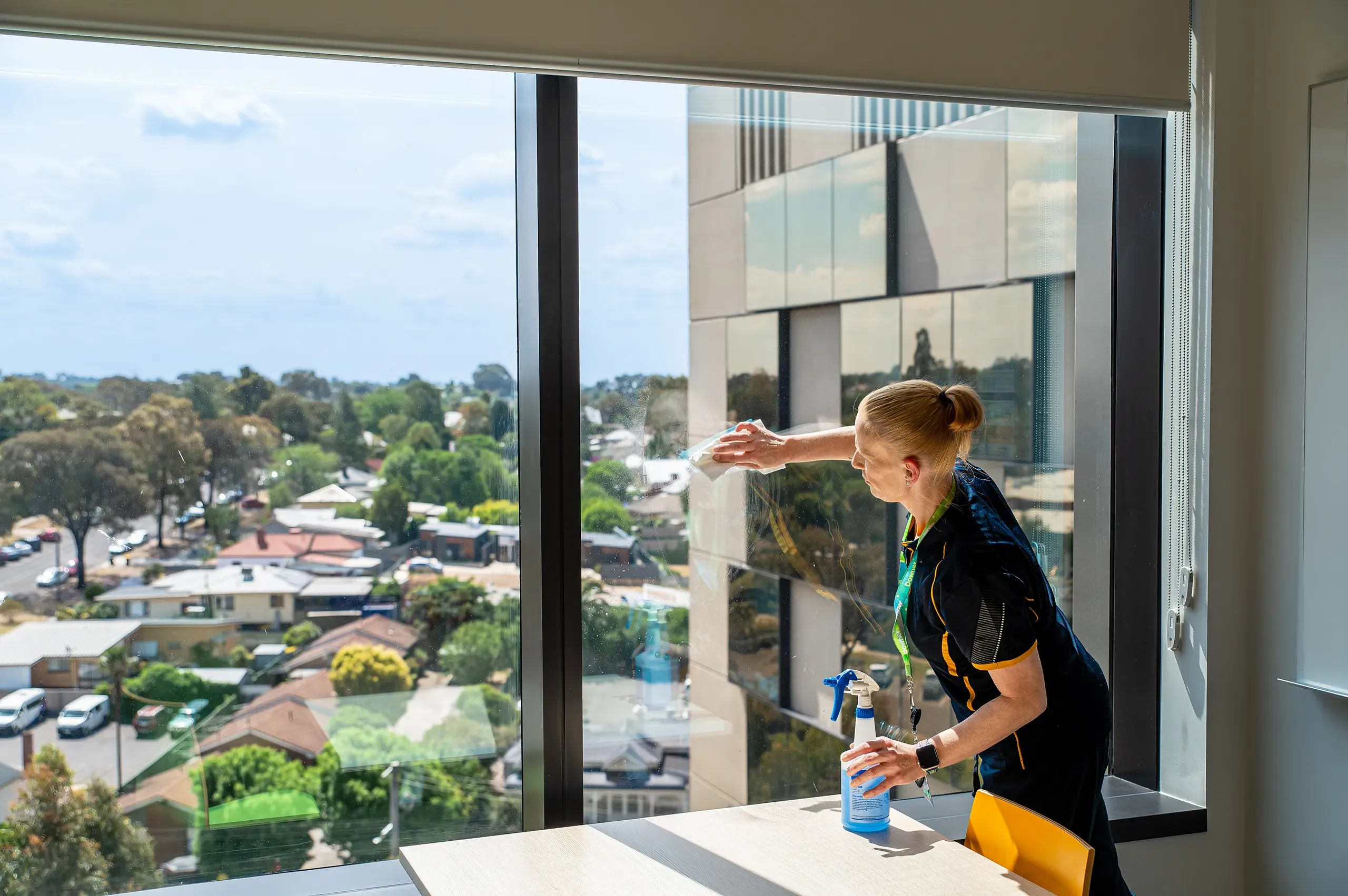 An indoor, eye-level shot shows a woman cleaning a window in a bright office. The woman, with her blonde hair in a bun, wears a dark blue shirt with yellow trim and a green lanyard. She holds a blue spray bottle and wipes the window with a white cloth. Through the window, a suburban neighborhood with houses, trees, and cars is visible under a blue sky with white clouds. A coloured wooden table and a yellow chair are in the foreground.